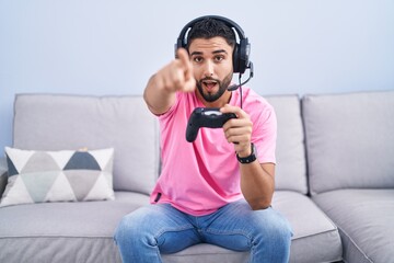 Hispanic young man playing video game holding controller sitting on the sofa pointing with finger surprised ahead, open mouth amazed expression, something on the front