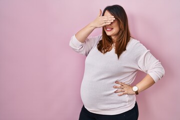 Pregnant woman standing over pink background smiling and laughing with hand on face covering eyes...