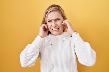 Young caucasian woman wearing white sweater over yellow background covering ears with fingers with...