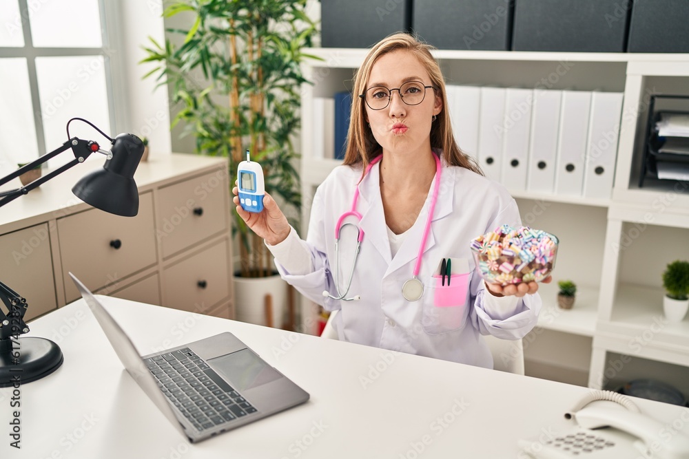 Poster Young blonde doctor woman holding glucometer and sweets looking at the camera blowing a kiss being lovely and sexy. love expression.