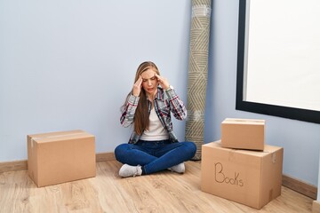 Young blonde woman sitting on the floor moving to a new home with hand on head for pain in head...