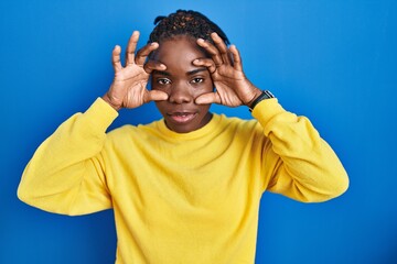 Beautiful black woman standing over blue background trying to open eyes with fingers, sleepy and tired for morning fatigue