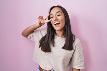 Young hispanic woman standing over pink background doing peace symbol with fingers over face, smiling cheerful showing victory