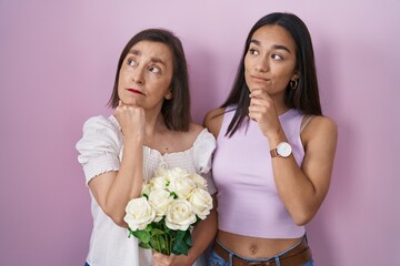 Hispanic mother and daughter holding bouquet of white flowers with hand on chin thinking about question, pensive expression. smiling with thoughtful face. doubt concept.