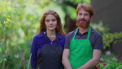 Two gardeners wearing aprons standing in green environment looking at camera in daylight. Horticulture entrepreneurs