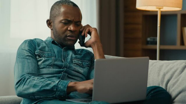 Serious african american man talking speaking phone typing on laptop at home ethnic businessman freelancer answering mobile call on couch working remote internet solving work issues using telephone