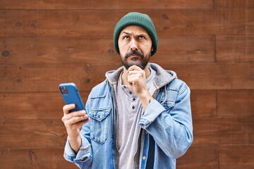 Young hispanic man using smartphone serious face thinking about question with hand on chin,...