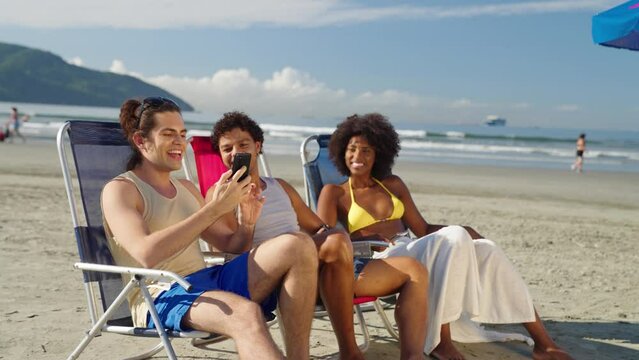 group of friends use smartphone to video call on the beach. Group of friends in swimwear enjoy talking at summertime. Travel, vacation and friendship concept.
