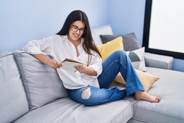 Fototapeta premium Young hispanic woman reading notebook sitting on sofa at home