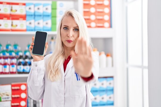 Caucasian woman working at pharmacy drugstore showing smartphone screen with open hand doing stop sign with serious and confident expression, defense gesture