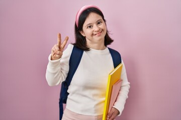 Woman with down syndrome wearing student backpack and holding books smiling looking to the camera showing fingers doing victory sign. number two.