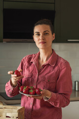 A smiling mature woman holds a strawberry berry next to a basket on the table in a bright kitchen. I cook jam, salad and eat fresh berries. Healthy eco-farm, organic homemade food