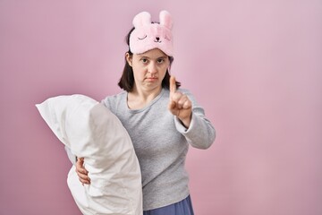 Woman with down syndrome wearing sleeping mask hugging pillow pointing with finger up and angry expression, showing no gesture