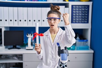 Teenager girl working at scientist laboratory holding degree annoyed and frustrated shouting with anger, yelling crazy with anger and hand raised