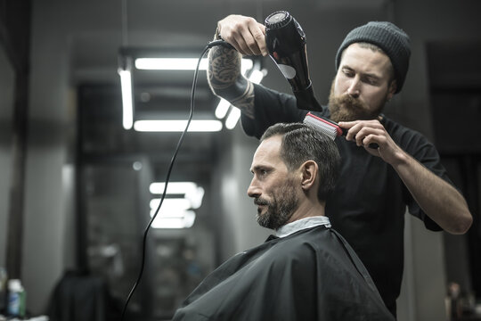 Unbelievable barber with a beard and a tattoo is drying the hair of his client in a black cutting hair cape in the barbershop. He is using a black hairdryer and a hairbrush. Horizontal.