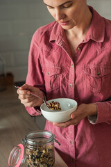 A mature woman puts homemade granola or muesli from a jar in a bowl. The concept of a healthy breakfast