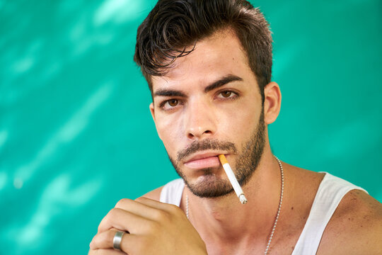 Real Cuban people and emotions, portrait of young hispanic man from Havana, Cuba looking at camera with serious expression, smoking cigarette and blowing smoke