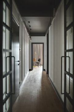 Low lighted corridor in a modern style with white walls and a parquet on the floor. There is an entrance door to a room with a wooden table, black chair and a trendy yellow lamp. Vertical.