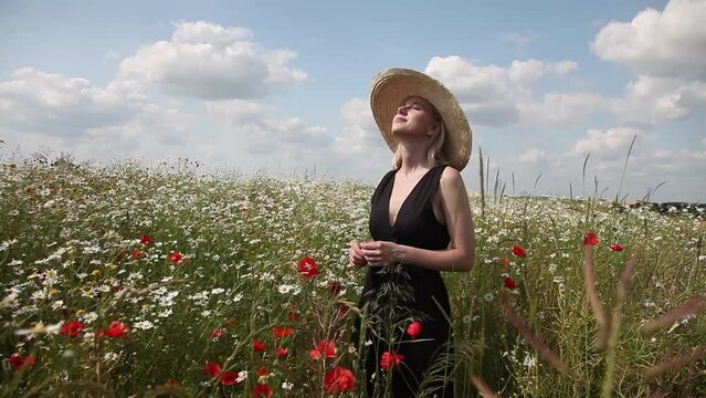 Stylish blonde woman in hat and black dress in chamomile meadow in sunny day with clouds on background