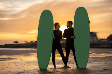 Happy millennial couple with surfboards standing on the beach on sunset time