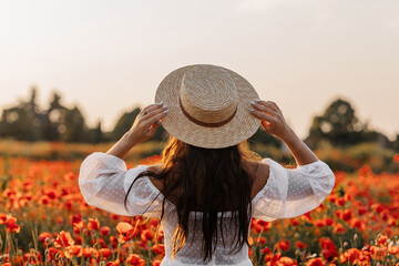 Beautiful young woman in short white dress and straw hat in field with poppies in evening at sunset...
