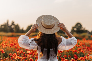 Beautiful young woman in short white dress and straw hat in field with poppies in evening at sunset and holds poppy in hand, Summer countryside nature flowers, Female relaxing, Rural simple life