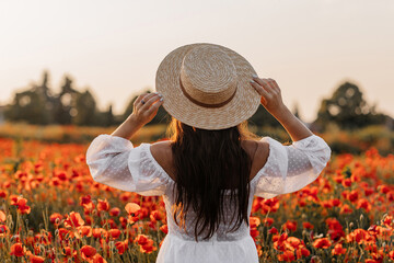 Beautiful young woman in short white dress and straw hat in field with poppies in evening at sunset and holds poppy in hand, Summer countryside nature flowers, Female relaxing, Rural simple life