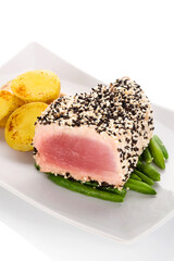 Grilled tuna steak wrapped in black and white sesame seeds on green beans with potatoes on white plate on white background. Culinary seafood eating.