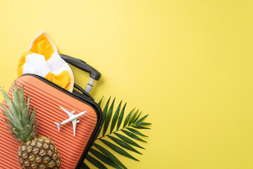 Summertime travel theme. Top view of orange suitcase, tiny airplane replica, beach gear, towel, ripe pineapple, palm leaves against a yellow background with empty space for text or advertisements © ActionGP