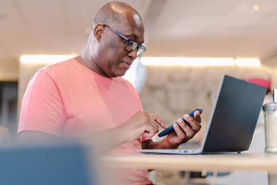 homem negro latino sentado na mesa de uma cafeteria com seu computador portatil usando seu celular para acessar a internet em uma cafeteria no Brasil