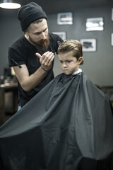 Trendy little boy in a black salon cape in the barbershop. Bearded barber with a tattoo in a black...