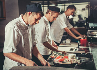 Three male cooks dressed in black aprons and hats preparing sushi set in the interior of modern...