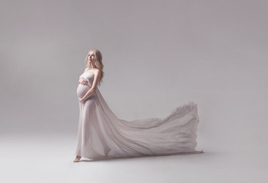 studio portrait of beautiful pregnant young woman in flying white dress with trail on light grey studio background