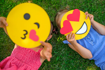 A child shows a kissing emoticon lying on the lawn grass in close-up. The girl hides behind a smile face in love