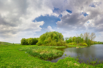 Spring landscape with lake, meadow on the coast and forest on the background of a sky with clouds in the afternoon