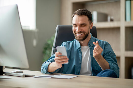Cheerful Businessman Making Finger Heart Gesture While Sitting At Desk In Office