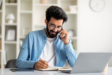 Young Indian Male Talking On Cellphone And Taking Notes To Notepad
