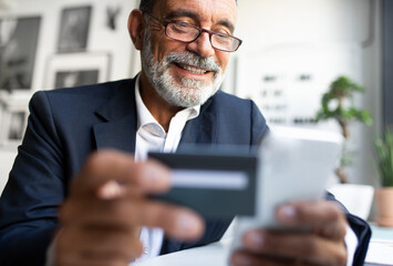 Cheerful mature european man in suit enjoy online shopping with phone and credit card in office