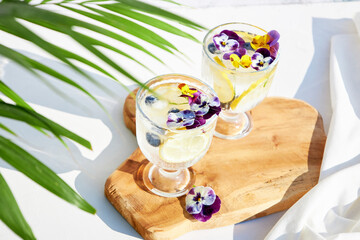 Iced soda with lemon and edible flowers under palm leaves on a bright sunny day. Summer cold cocktails and drinks. Detox water.