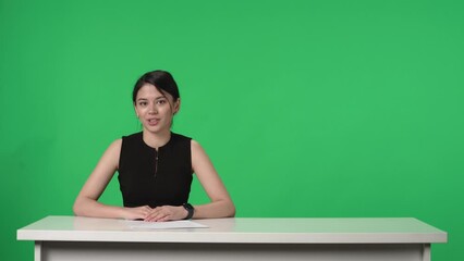 Reporting of a leading Asian woman, sit at a table in front of a green screen. The TV presenter speaks confidently and points towards the green screen. Advertising area, workspace mock up.