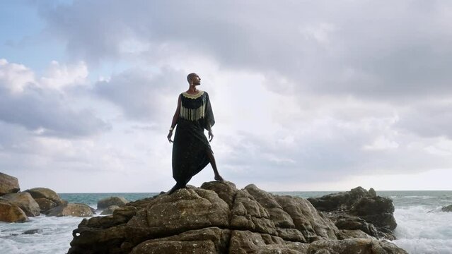 LGBTQIA black person in boutique dress, jewelry stands on rock on stunning ocean coastline skyline. Gender fluid ethnic divine model in luxury outfit fluttering in the wind against dramatic sky.
