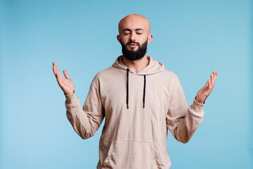 Fototapeta premium Arab man praying and making worship gesture with open arms and closed eyes. Young religious person praising, pleading to god with raised hands and asking for forgiveness concept