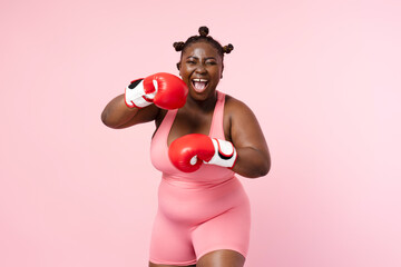 Positive african american woman wearing sports uniform red boxing gloves isolated on pink background