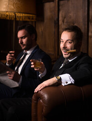 Happy rich men spending free time in restaurant. Luxury men sitting on sofa, drinking whiskey and smoking cigars.