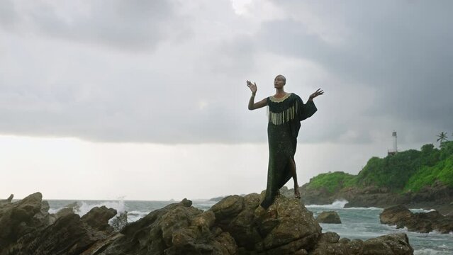 LGBTQ bipoc person in luxury dress, boutique golden jewelry poses against lighthouse on cliff, ocean, looks aside. Divine gender fluid multiethnic model in epic outfit stands on rock. Cloudy sky.