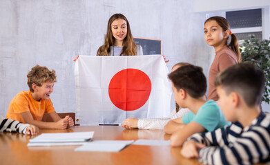 Naklejka na ściany i meble Smiling young female teacher explaining culture Japan while showing national flag to schoolchildren preteens in classroom