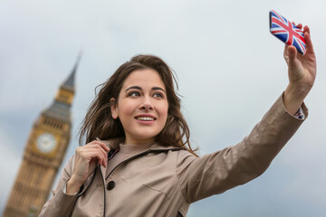 Girl or young woman tourist on vacation taking a selfie photograph by Big Ben with Union Jack cell...