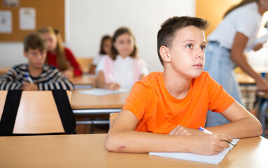 Young boy sitting at desk in classroom and writing in test book.