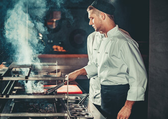 Young white chef in black apron and hat standing near the brazier whith coals. Man cooking beef steak in the interior of modern professional kitchen