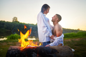 Happy Couple relaxin, having fun and hugs with fire in camping on nature near water of river or lake in summer sunny evening in sunset. Family or lovers have date and rest outdoors. Concept of love